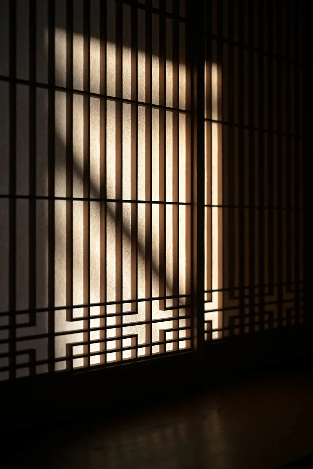 Sun through a wooden lattice lays bars of light across a paper screen.