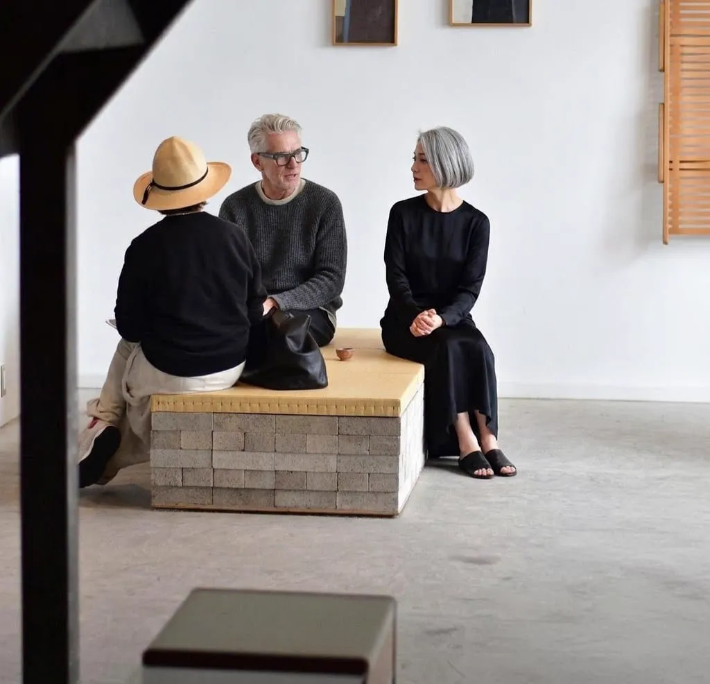 Three people talking on a low bench in a pale room.